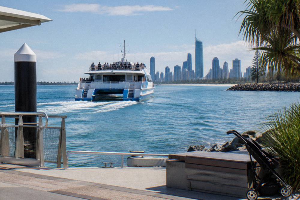A large power catamaran cruising past a city skyline with passengers along the upper rail