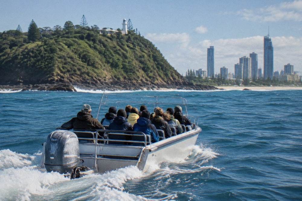A small open safari-style boat with a dozen passengers moving across open ocean near a headland